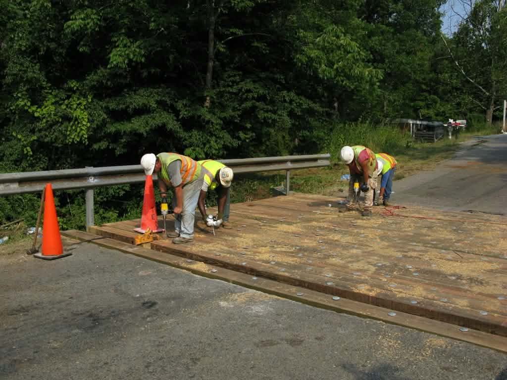 Four workers in hard hats and safety vests kneel on fresh wooden bridge decking beside orange cones.