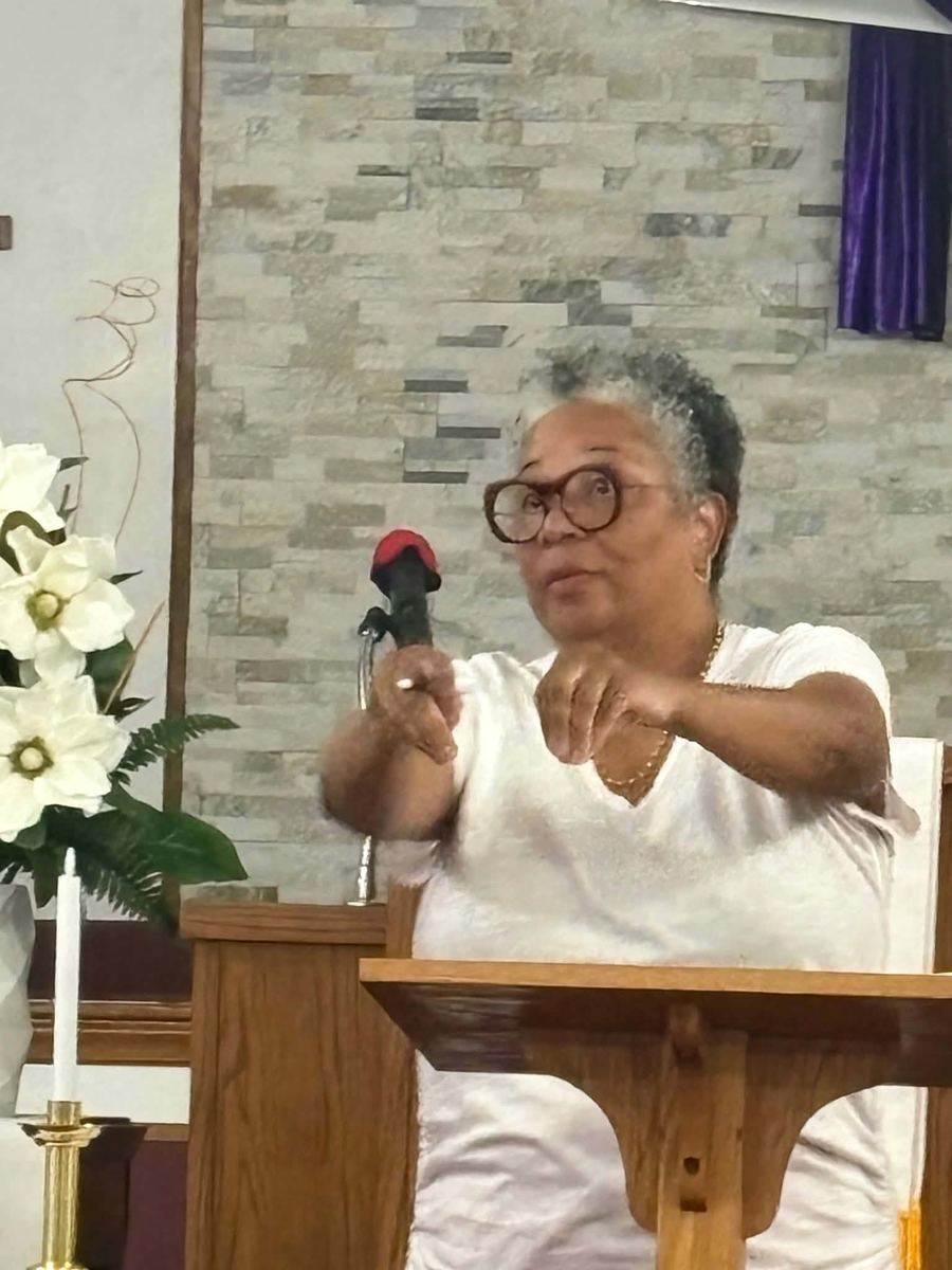 A woman in glasses speaks and gestures from behind a wooden lectern with a microphone.